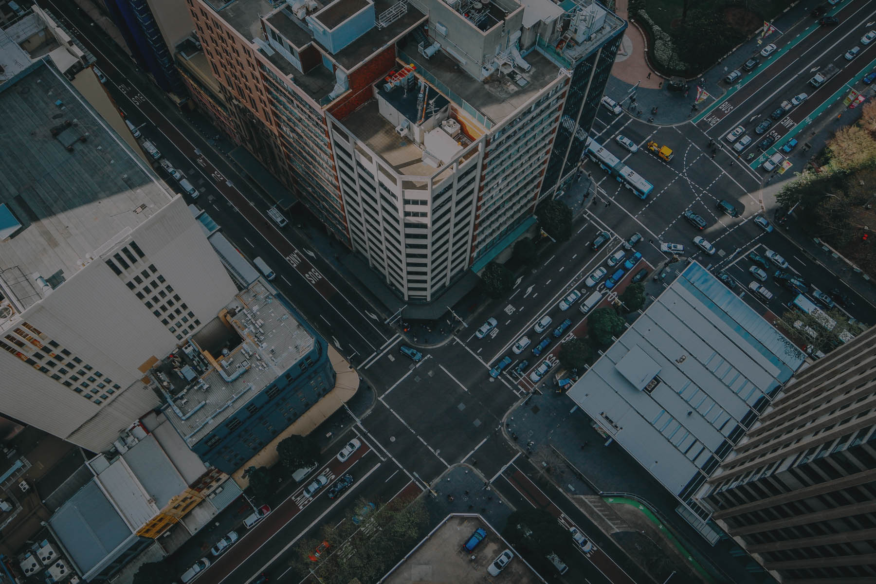 Aerial view of a city intersection with traffic, buildings, and rooftops; dark blue and gray tones, urban busy mood.